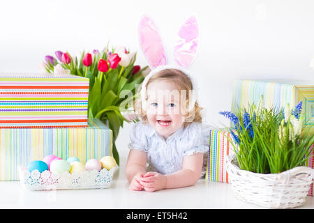 Happy laughing toddler girl wearing a blue dress et oreilles de lapin de Pâques avec des oeufs colorés, présente et fleurs de printemps Banque D'Images