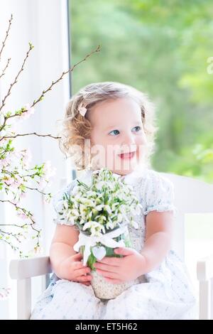 Tout-petit curly belle jeune fille en robe blanche assise dans une chaise à bascule blanc holding premier printemps fleurs dans vase de cristal Banque D'Images