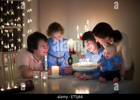 Les jeunes professionnels famille avec trois enfants célébration de l'anniversaire de leur fils souffler les bougies d'un gâteau dans une sombre salle de séjour Banque D'Images