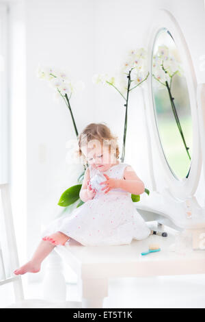 Heureux bébé fille avec des cheveux bouclés vêtue d'une robe blanche à jouer avec le maquillage et les cosmétiques en face d'un miroir rond Banque D'Images