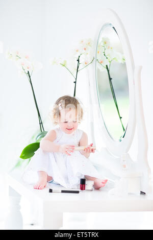 Heureux bébé fille avec des cheveux bouclés vêtue d'une robe blanche à jouer avec le maquillage et les cosmétiques en face d'un miroir rond Banque D'Images