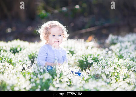 Belle curly bébé fille jouant avec les premières fleurs du printemps dans un magnifique parc ensoleillé de printemps Banque D'Images