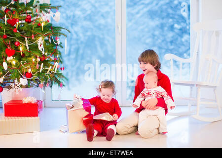 Trois enfants d'ouvrir des cadeaux de Noël, profitant de matin de Noël dans une salle de séjour avec un arbre décoré et fenêtre dans un jardin enneigé Banque D'Images