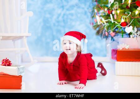 Cute little girl bouclés dans une robe rouge et Santa hat jouer sous un arbre de Noël avec présente assis sur le plancher Banque D'Images