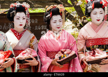 Japon, Kyoto. Trois geisha japonais, geiko, portant des kimono roses et crème, posant dans une rangée à l'extérieur dans le soleil printanier par quelques arbustes verts. Banque D'Images