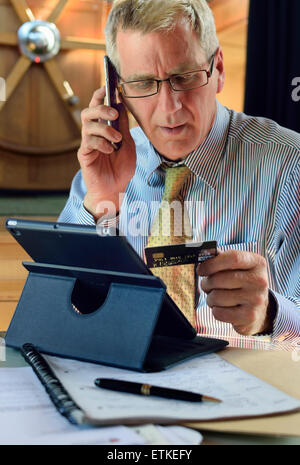 Man & iPad avec smartphone iPhone dans son bureau de péniche effectuer une transaction de carte bancaire en ligne sécurité de lecture informations au dos de la carte Banque D'Images