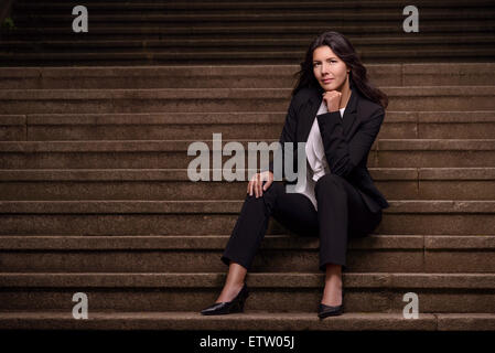 Smiling woman élégant dans un costume sombre slack assis sur un vol de mesures concrètes avec sa main posée sur sa joue à la th Banque D'Images