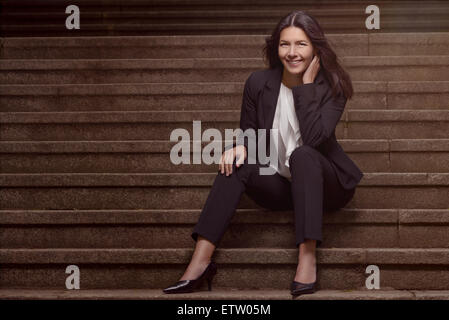 Smiling woman élégant dans un costume sombre slack assis sur un vol de mesures concrètes avec sa main posée sur sa joue à la th Banque D'Images