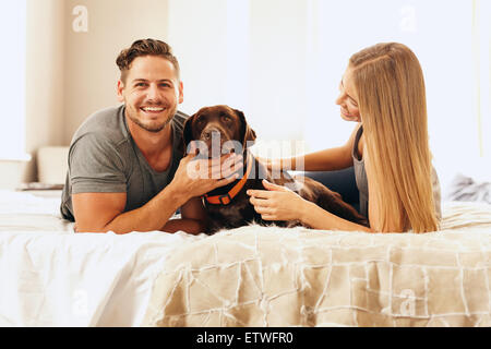 Portrait of happy young couple on bed with dog in matin. L'homme de caresser le chien et souriant. Banque D'Images