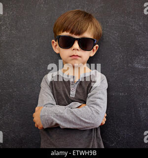 Portrait of little boy wearing sunglasses looking at camera, les bras croisés. Petit enfant devant un tableau noir qui pose pour Banque D'Images