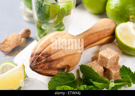 Centrifugeuse en bois et les ingrédients pour faire des mojitos. La glace, feuilles de menthe fraîche, citron vert et sucre oursons sur une planche à découper en verre blanc Banque D'Images