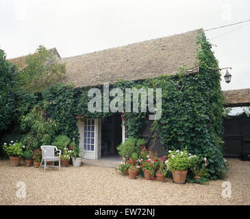 Chaise en osier blanc et de pots de plantes à fleurs d'été sur le gravier terrasse devant de maison de campagne française avec des portes ouvertes Banque D'Images