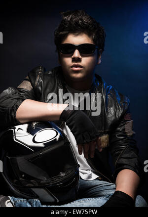 Portrait de joli garçon portant des lunettes de soleil, veste en cuir élégant et holding helmet posant dans le studio sur fond bleu Banque D'Images