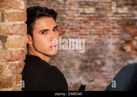 Beau jeune homme assis contre le mur de briques, looking at camera Banque D'Images