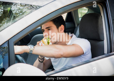 Beau jeune homme conduisant sa voiture tout en mangeant de la nourriture dans le trafic Banque D'Images