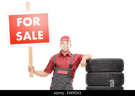 Cheerful male mechanic standing par une pile de pneus et la tenue d'un big red for sale sign isolé sur fond blanc Banque D'Images