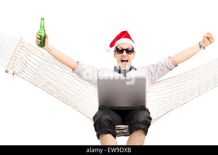 Joyful guy with Santa hat travaillant sur un ordinateur portable assis dans un hamac et boire de la bière isolated on white Banque D'Images