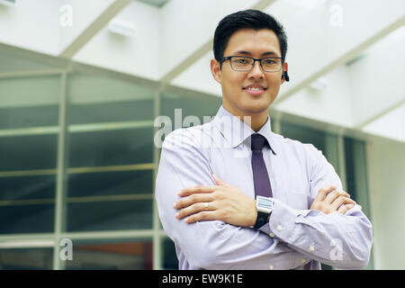 Portrait of smiling asian businessman. L'homme se trouve dans une rue à l'égard des immeubles de bureaux et les bras des croix looking at camera Banque D'Images