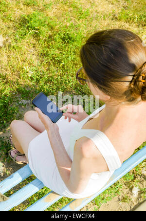 A senior woman est assise sur un banc de Woden et utilisé un smartphone pour envoyer un message texte Banque D'Images