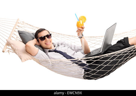 Jeune homme couché dans un hamac avec un ordinateur portable sur ses genoux et boire un cocktail orange isolé sur fond blanc Banque D'Images