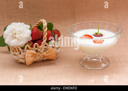 Yaourt au fraise au bol en verre sur fond beige et un sac, panier de fraises fraîches, feuilles vertes et roses Banque D'Images