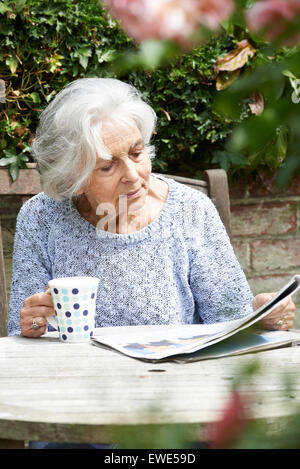 Senior Woman Relaxing In Garden Reading Newspaper Banque D'Images