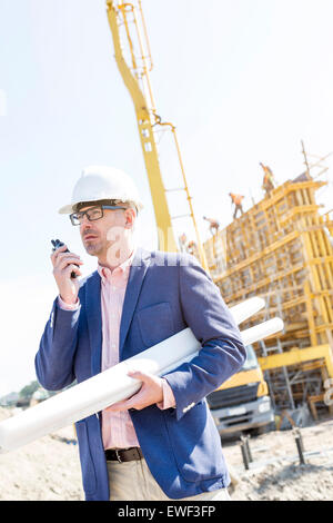 Superviseur en utilisant talkie-walkie tandis que holding blueprints at construction site Banque D'Images