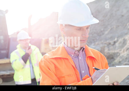 Engineer writing on clipboard at construction site with colleague in background Banque D'Images