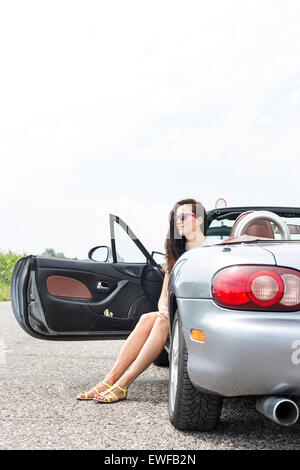 Thoughtful woman sitting in convertible sur route de campagne contre le ciel clair Banque D'Images