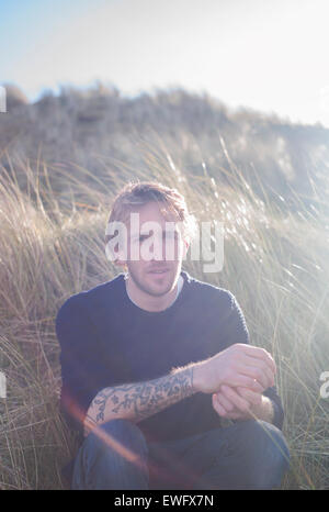 Portrait d'un homme avec un tatouage assis dans les dunes de sable à la plage en été. Banque D'Images