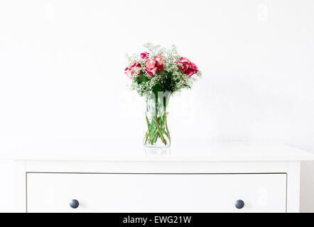 Cette image montre un arrangement floral minimaliste avec des roses rouges et blanches, complétées par le souffle de bébé (Gypsophila paniculata) dans un vase simple. L'arrangement est placé sur un tiroir coffre avec un fond blanc propre, soulignant l'élégance et la simplicité. Banque D'Images