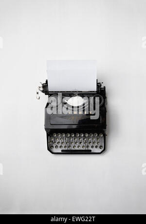 Une machine à écrire vintage repose sur un fond blanc, accompagnée d’une feuille de papier, évoquant l’essence du journalisme traditionnel. La composition en noir et blanc souligne le design classique et ancien et la valeur nostalgique de la machine à écrire. Banque D'Images