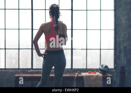 Une femme forte, d'athlétisme est debout regardant par la fenêtre dans une salle de sport city loft. Une serviette est jeté par-dessus son épaule, et sa main est sur sa hanche. Banque D'Images