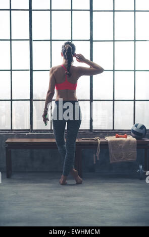 Un ajustement, fort, musclé femme se tient debout, les jambes croisées, tout en regardant par la fenêtre. Dans une main, elle tient son appareil, écouter de la musique. De l'autre main, elle est à l'écouteur. Banque D'Images