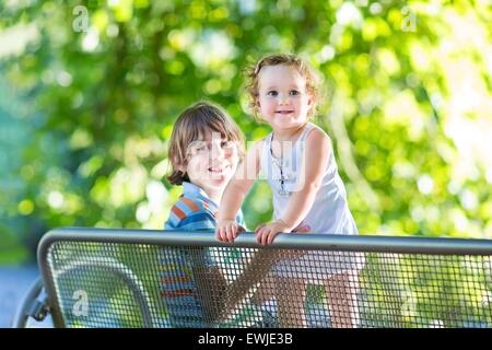 Adorable bébé fille aux cheveux bouclés portant une robe bleue jouant avec son frère assis sur un banc dans un parc à l'été ensoleillé Banque D'Images