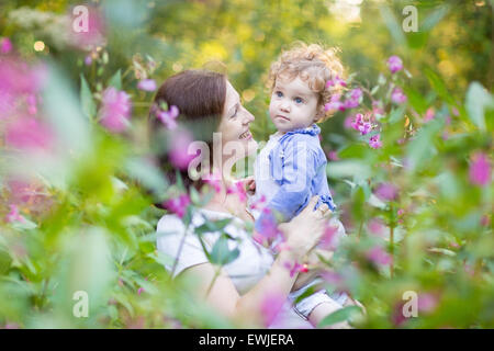 Young pregnant mother holding her baby fille dans un jardin au coucher du soleil parmi les magnifiques fleurs roses Banque D'Images