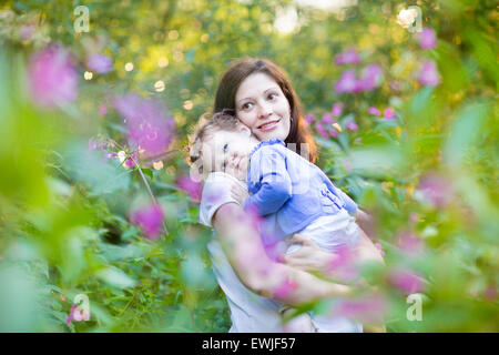 Young pregnant mother holding her baby daughter fatigué dans un parc avec de belles fleurs roses et mauves au coucher du soleil Banque D'Images