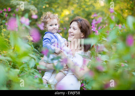 Young pregnant mother holding her baby fille dans un jardin au coucher du soleil parmi les magnifiques fleurs roses Banque D'Images