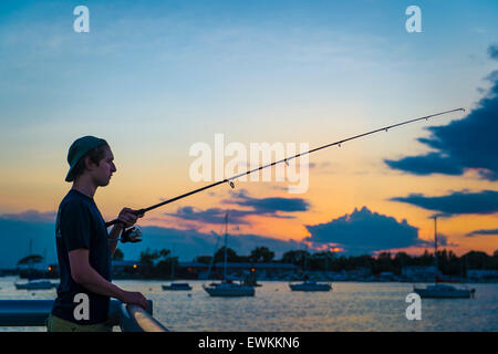 Port Washington, New York, USA. 26 juin 2015. Un jeune homme, vêtu de l'arrière de la PAC, est la pêche au coucher du soleil, les approches sur le quai de la ville sur les rives de Manhasset Bay dans le village de la rive nord de Long Island Gold Coast. Credit : Ann E Parry/Alamy Live News Banque D'Images