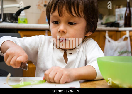 Caucasien enfant, garçon, 4-5 ans, assis à l'intérieur à table de cuisine et de couper l'alimentation avec un couteau sur la planche à hacher avec expression concentrée. Banque D'Images