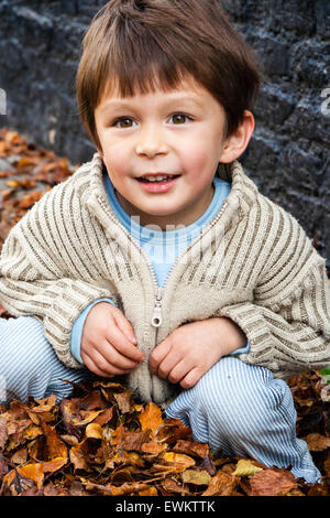 Caucasien enfant, garçon, 5-6 ans, à l'extérieur par un mur de brique peint en noir, accroupi entre brown l'automne les feuilles tombées, looking up and smiling. Banque D'Images