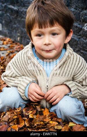 Caucasien enfant, garçon, 5-6 ans, à l'extérieur par un mur de brique peint en noir, accroupi entre brown automne les feuilles tombées, regardant vers le bas de façon réfléchie. Banque D'Images