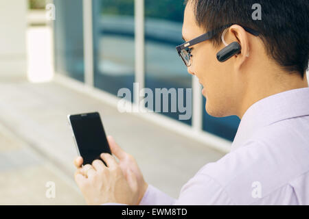Jeune homme d'affaires chinois de taper votre numéro de téléphone sur le smartphone et de parler avec casque bluetooth device Banque D'Images