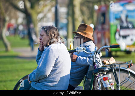 Un homme noir et une femme blanche s'asseoir sur un banc dans un parc de Londres pour regarder les gens dans la rue alors qu'elle a une cigarette Banque D'Images