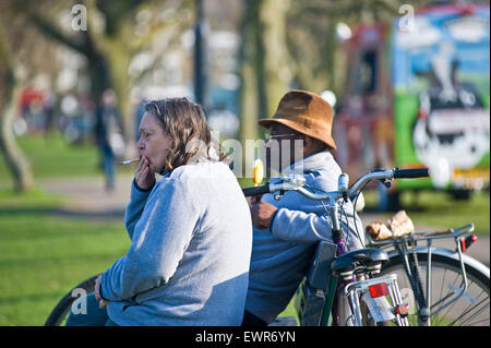 Un homme noir et une femme blanche s'asseoir sur un banc dans un parc de Londres pour regarder les gens dans la rue alors qu'elle a une cigarette Banque D'Images