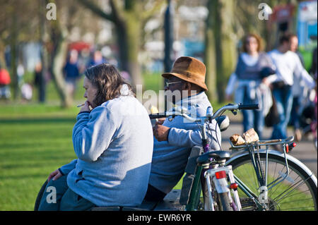 Un homme noir et une femme blanche s'asseoir sur un banc dans un parc de Londres pour regarder les gens dans la rue alors qu'elle a une cigarette Banque D'Images