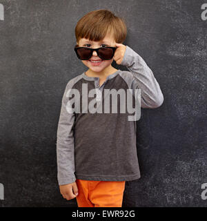 Portrait of happy little boy wearing sunglasses smiling at camera. Petit boy peering over ses lunettes contre tableau noir. Banque D'Images