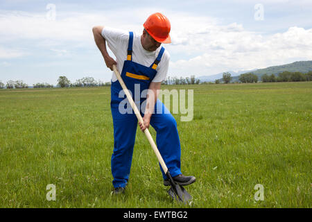L'homme dans les vêtements de travail et casque, est de creuser avec une pelle, dans une pelouse verte. Il s'agit d'une journée ensoleillée en été. Le travailleur est en Banque D'Images