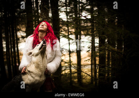 La substance des rêves et cauchemars : Une jeune femme aux cheveux rouge caucasian girl alone dans une sombre forêt de pins avec un chien-loup blanc comme Banque D'Images