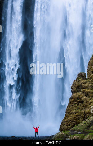 L'homme avec son bras en l'air debout au pied de la cascade de Skogafoss, Islande Banque D'Images